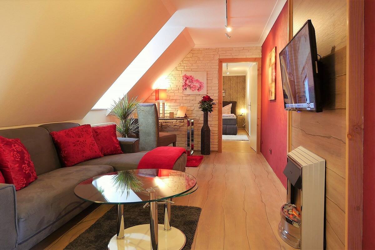Ein modernes Wohnzimmer mit Glastisch, grauem Sofa mit roten Kissen, einer Topfpflanze und einem Kamin.