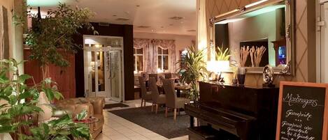 Un vestíbulo de hotel con un piano, zona de comedor y un cartel con opciones de menú.