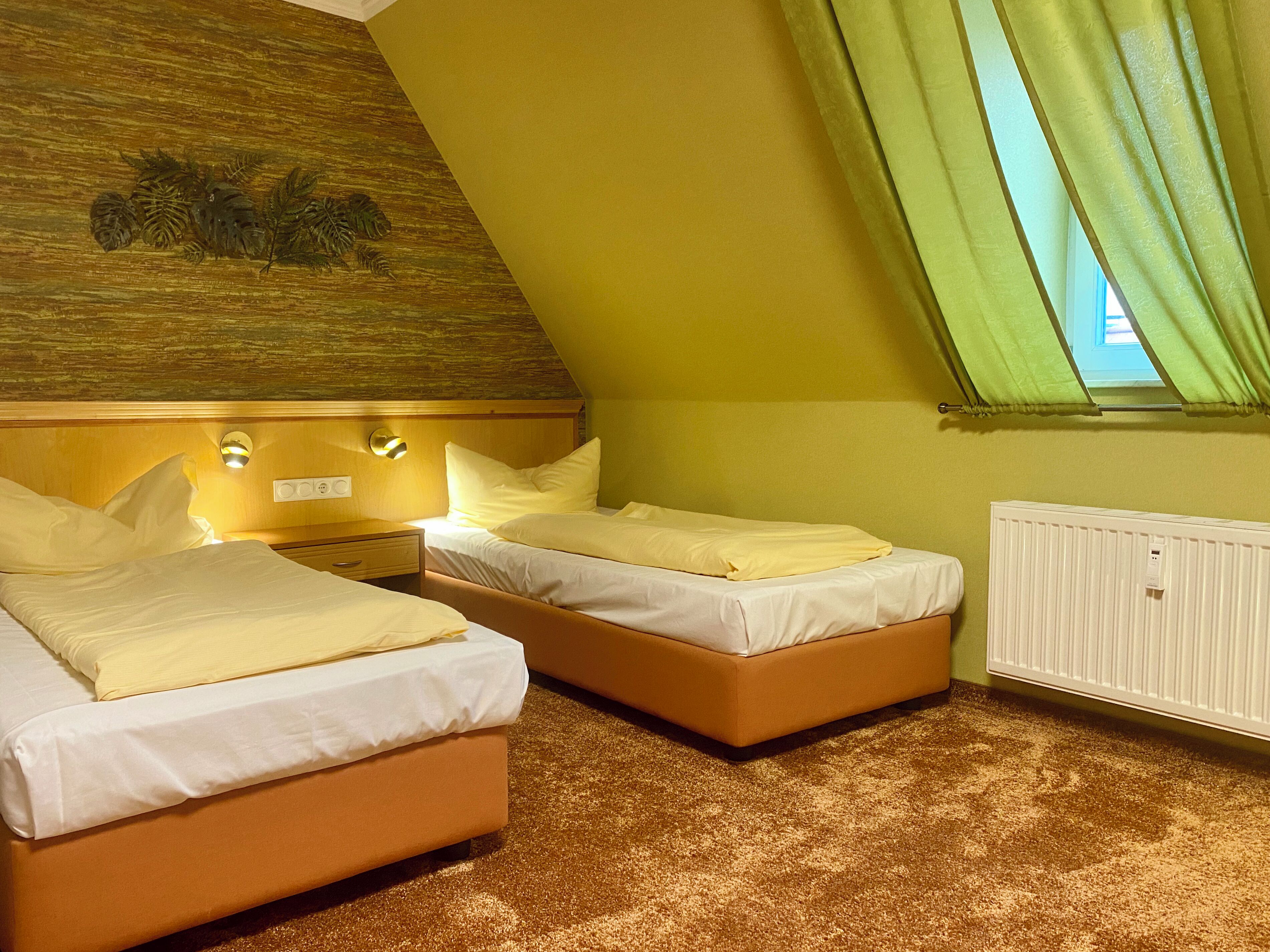Una habitación con dos camas, cabecera de madera, ventanal con cortinas verdes y un radiador.