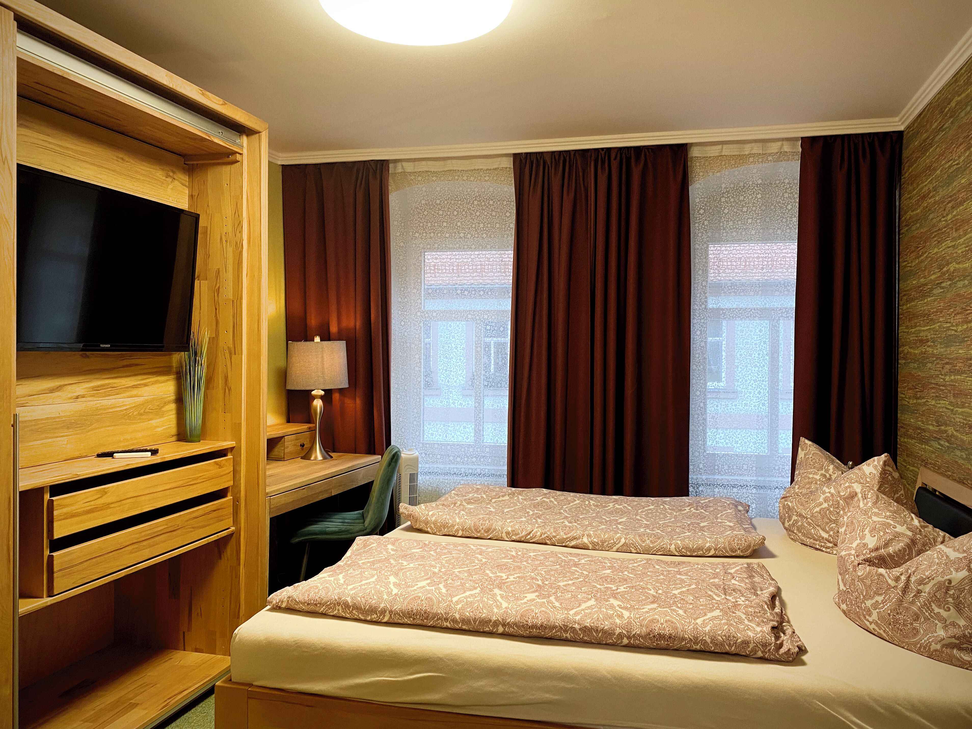 Habitación de hotel con televisor, escritorio, dos camas y ventanas amplias con cortinas.