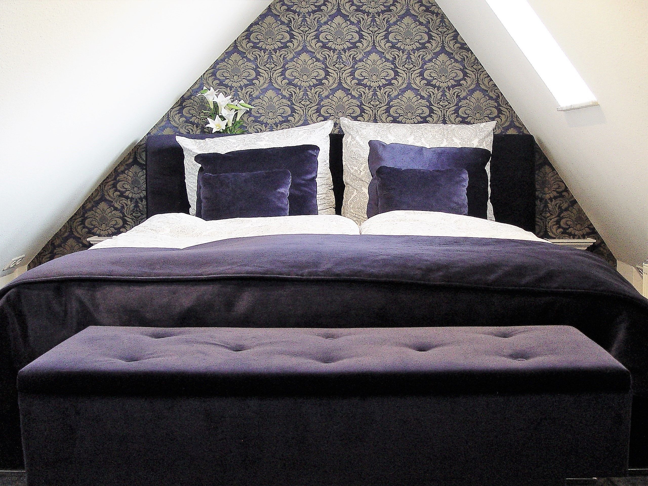 A bed with a dark purple headboard and pillows, a white lace pillow, and a dark purple bench in front.