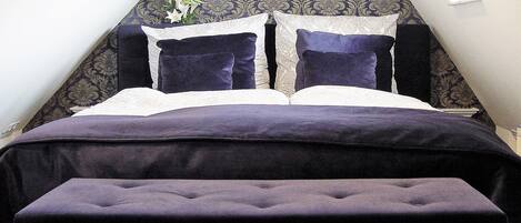 A bed with a dark purple headboard and pillows, a white lace pillow, and a dark purple bench in front.