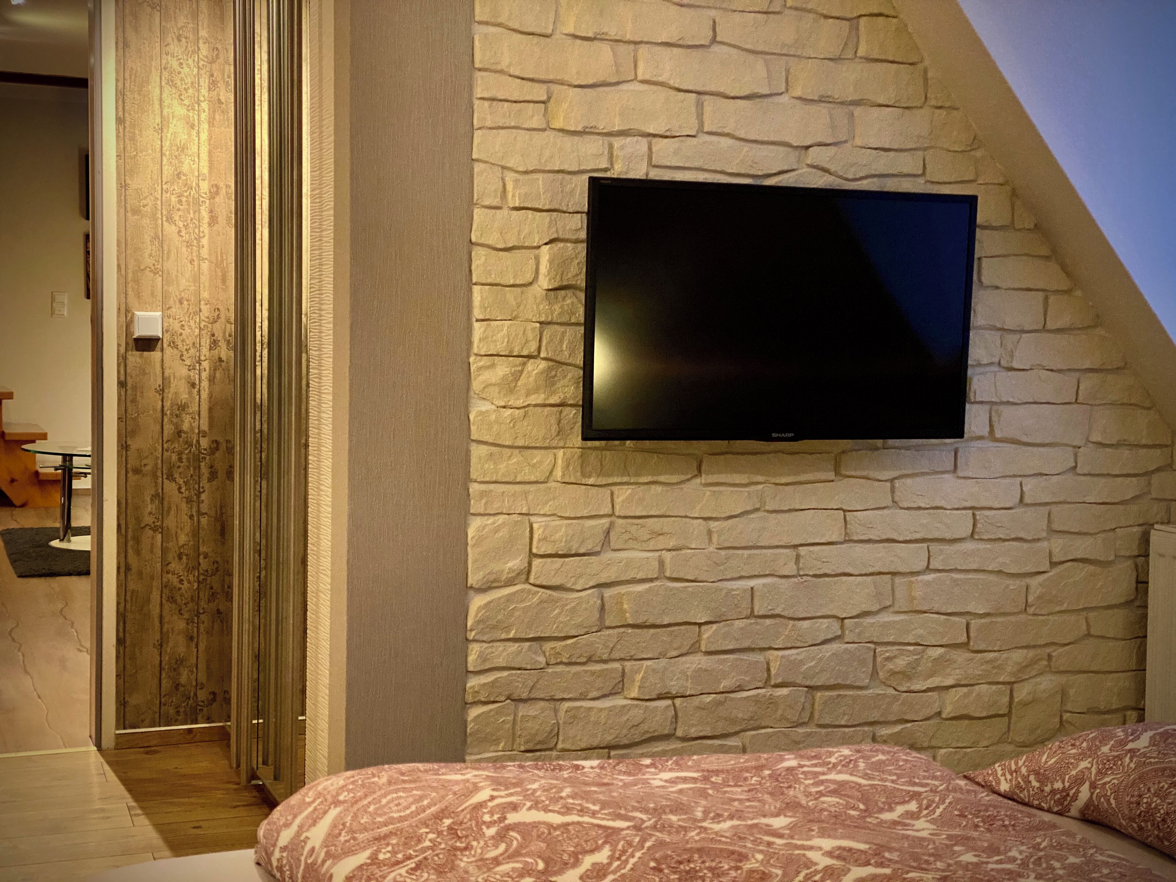 Quarto com parede de pedra, TV de tela plana fixada na parede, cama com roupa de cama estampada e porta de madeira que dá para outro cômodo.