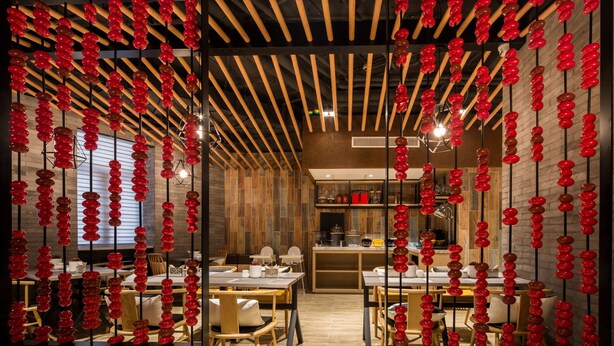 A modern restaurant with wooden walls and ceiling, red hanging decorations, and a central kitchen area.