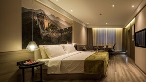 A hotel room with a large mural of the Great Wall of China, a bed with a green throw, a desk with a chair, and a TV on the wall.
