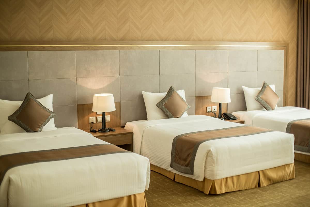 A hotel room with three beds, each with a headboard, bedside tables with lamps, and a large wall panel behind the beds.