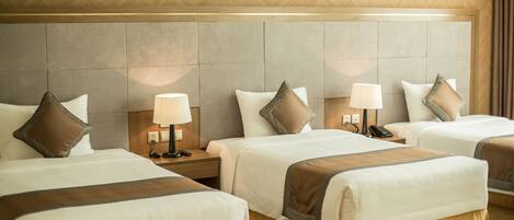A hotel room with three beds, each with a headboard, bedside tables with lamps, and a large wall panel behind the beds.