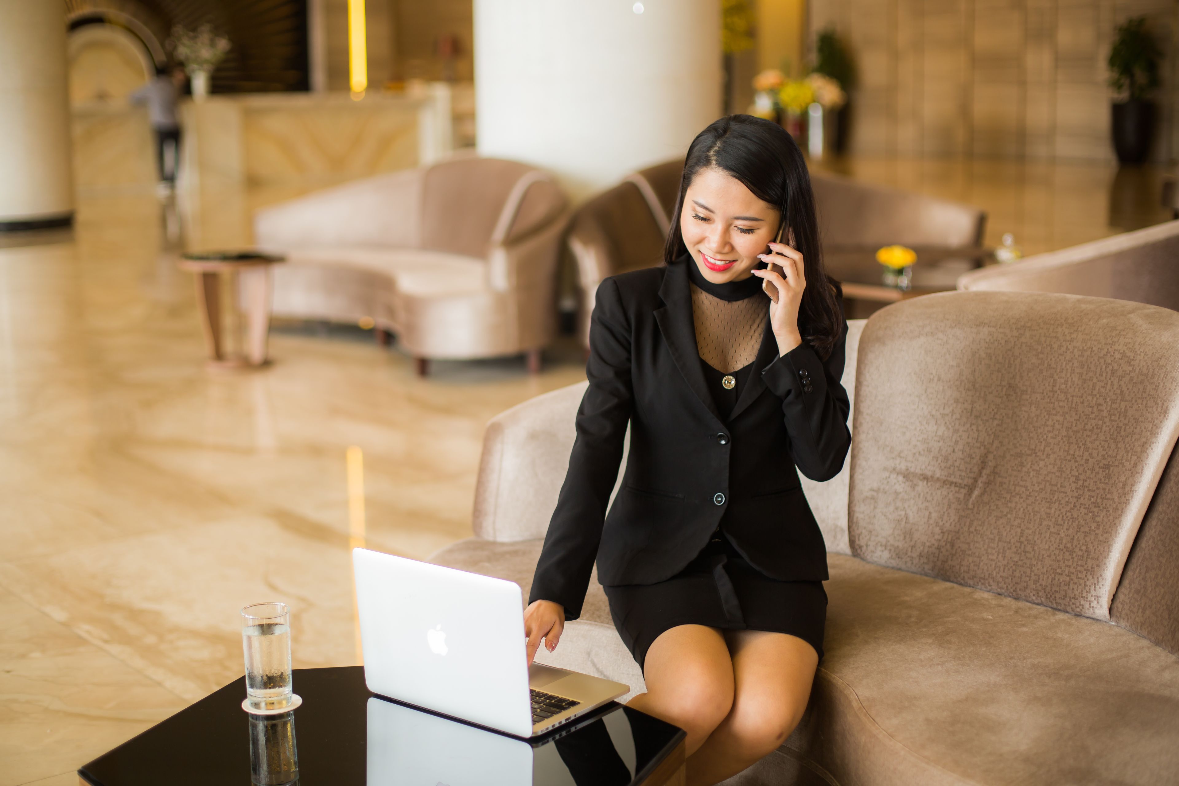 A woman sitting on a beige sofa in a modern lobby with a laptop on a small table.