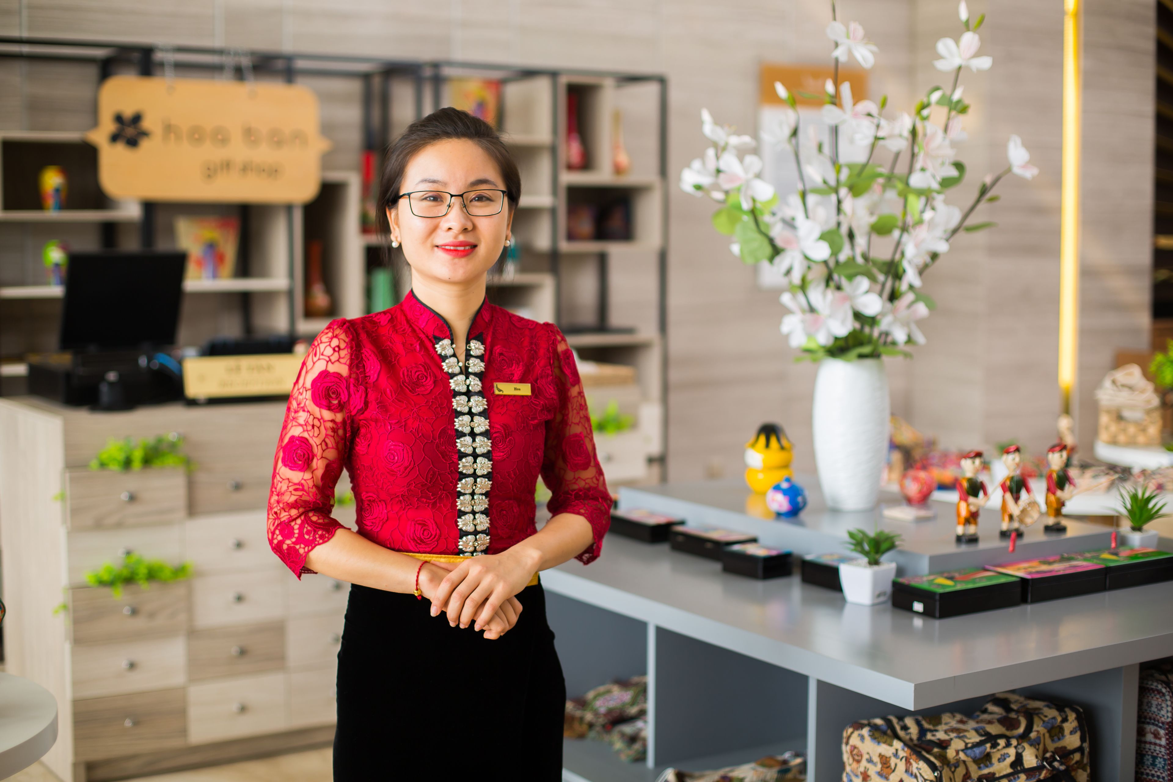 A woman in a red lace blouse and black skirt standing in a modern room with wooden drawers, a white vase with flowers, and a sign that reads "hoen gift shop".