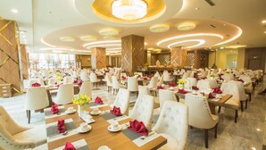 Ampla sala de banquetes com mesas redondas preparadas para refeições, com cadeiras brancas e guardanapos vermelhos.