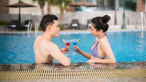 Um casal sentado perto de uma piscina, com bebidas, cercado por espreguiçadeiras e guarda-sóis.