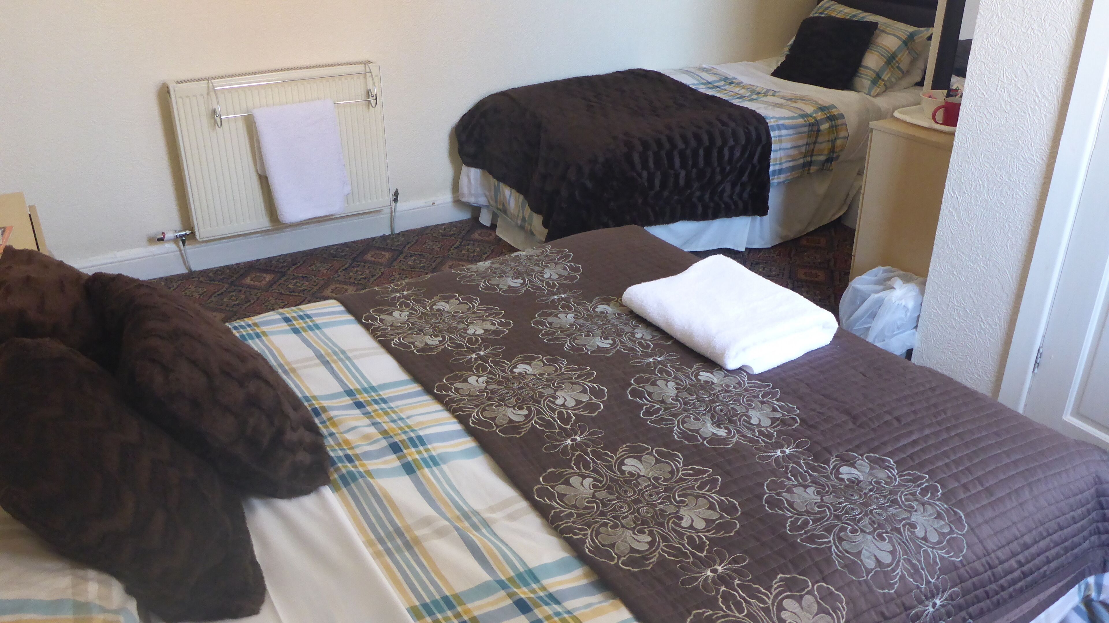 A hotel room with two beds, a radiator, and a bedside table with a cup.