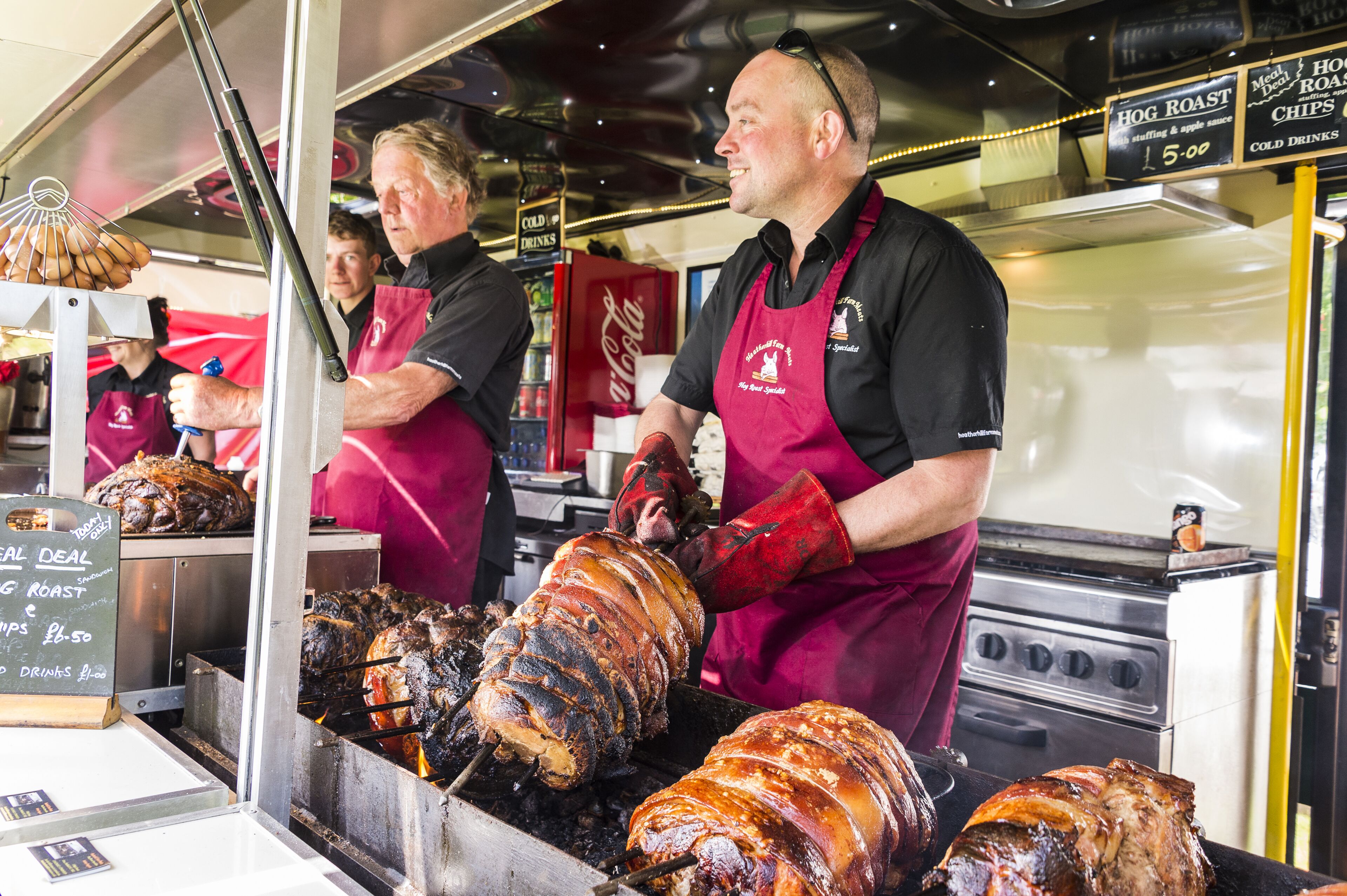 A food stall with a large spit-roasted pig, staff wearing red aprons, and a Coca-Cola cooler in the background.