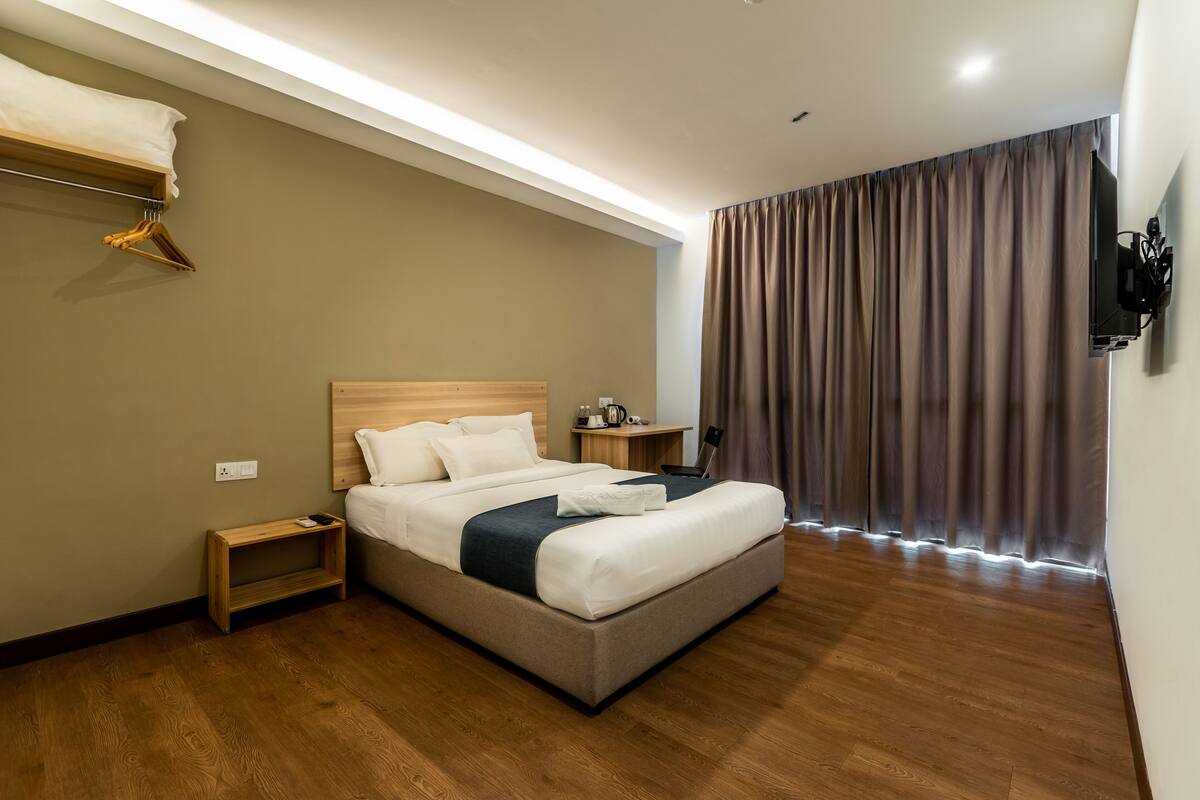 A modern hotel room with a large bed, wooden floor, a bedside table, a small shelf, and a wall-mounted TV.