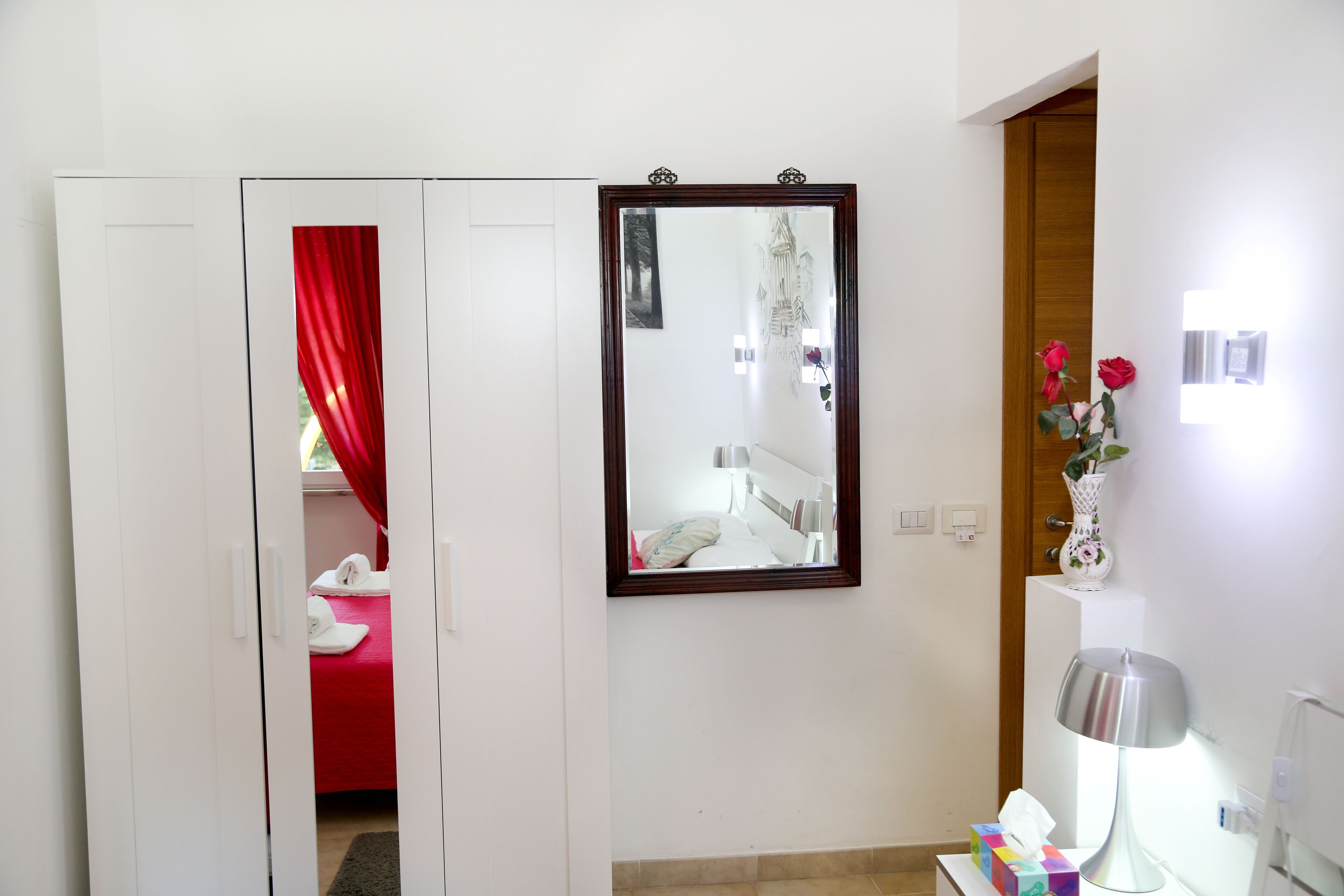 A hotel room with a bed, a mirror, a bedside table with a lamp, and a vase with flowers.