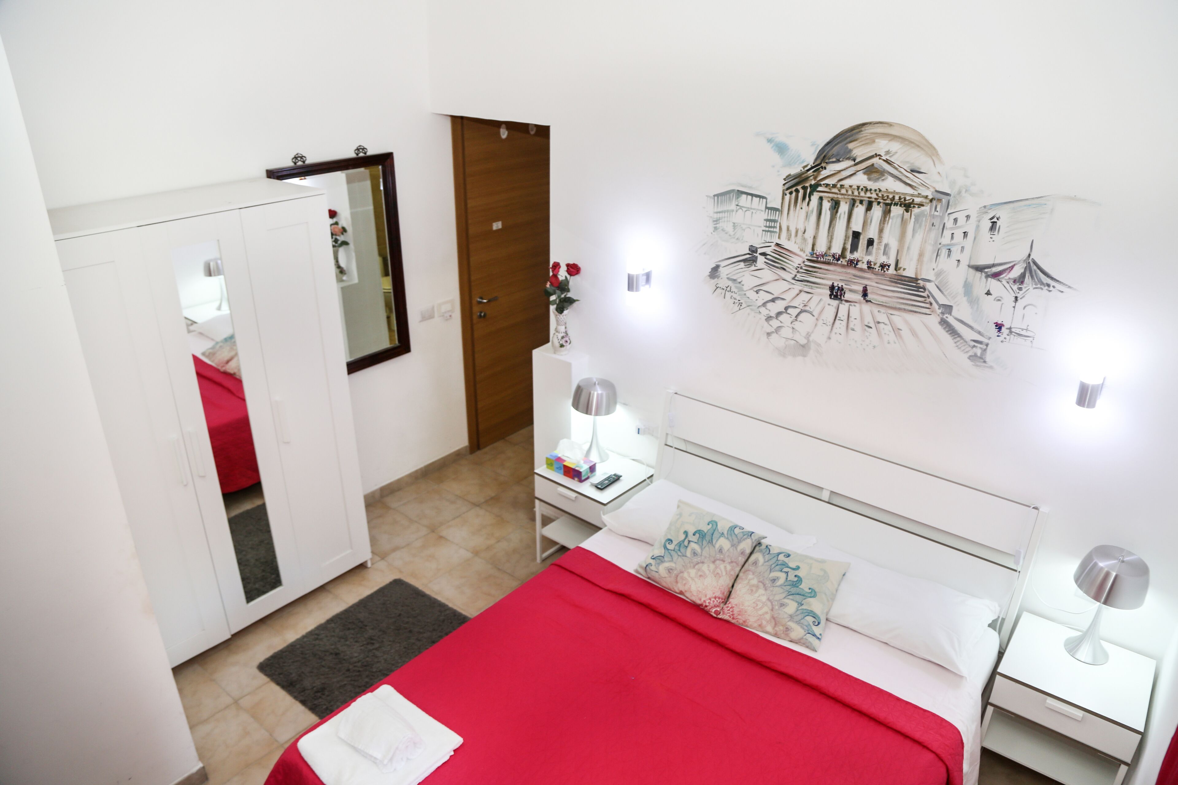 A bedroom with a red bedspread, white headboard, bedside tables with lamps, a wardrobe, and a wall mural.
