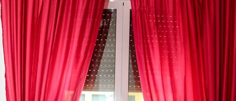 Una habitación con cama, ventana con cortinas rojas y vista a un edificio.