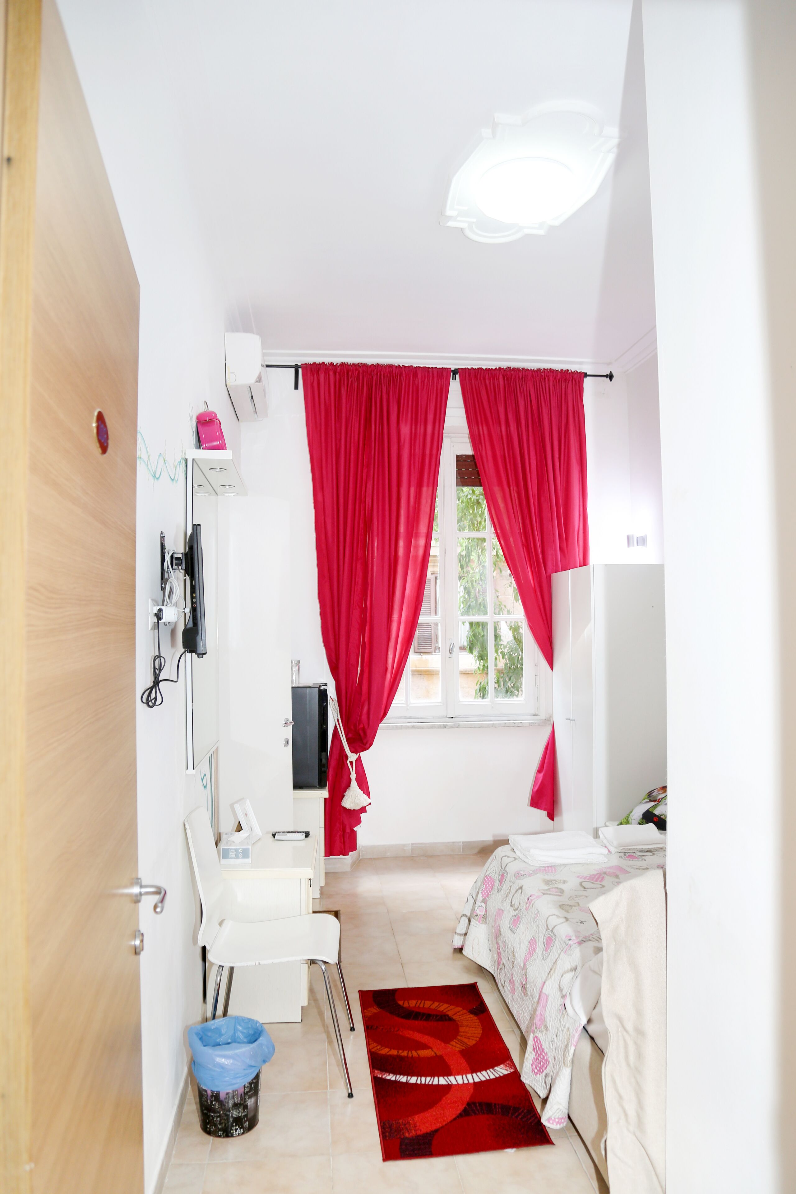 Une chambre à coucher avec un lit, un bureau, une chaise, une fenêtre avec des rideaux rouges et une petite poubelle.