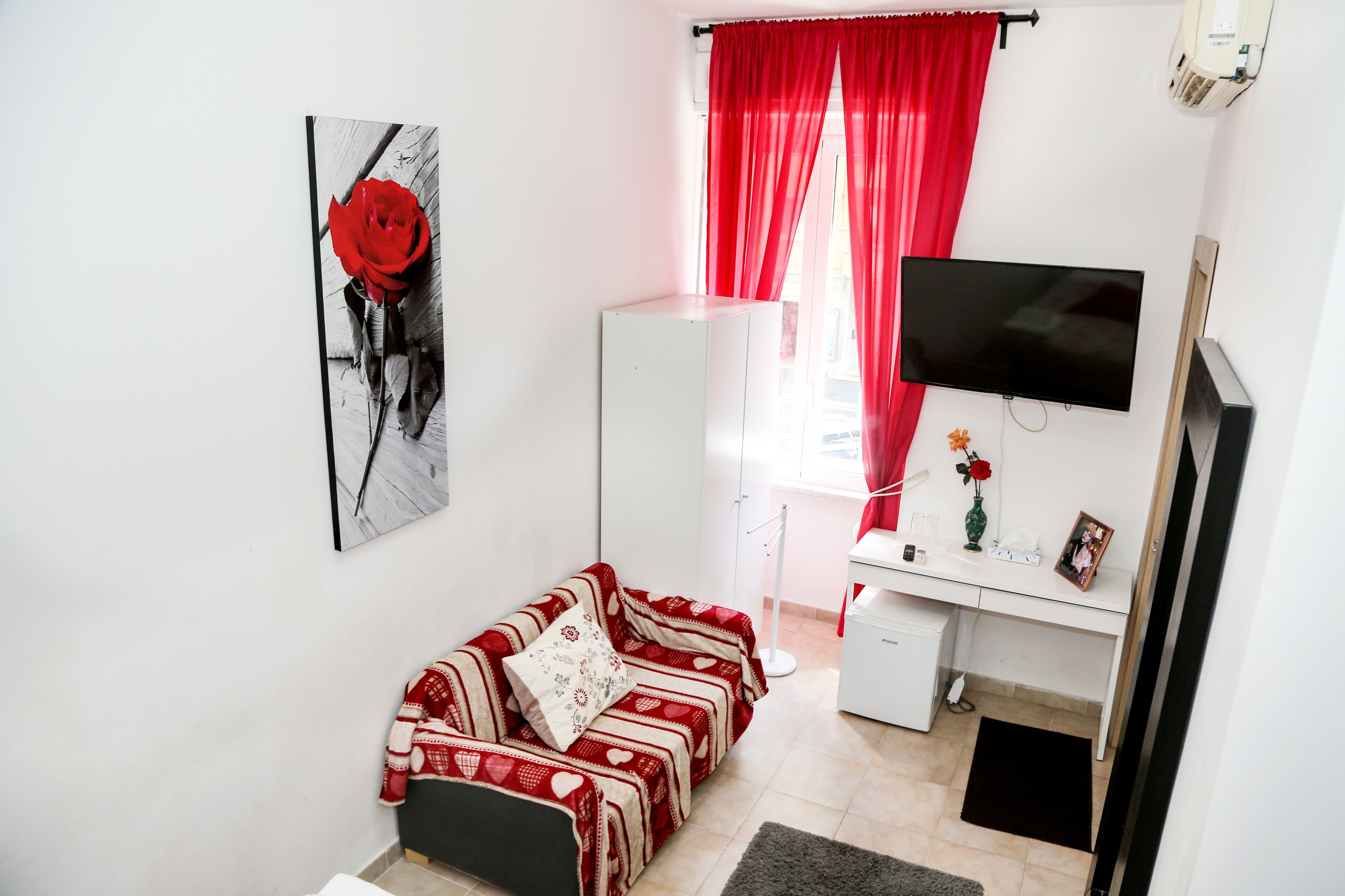 Un dormitorio con cama, escritorio, televisión y ventana con cortinas rojas.