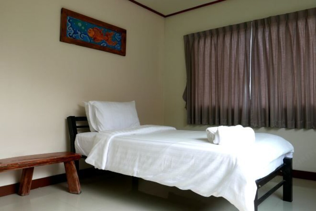 A single bed with a white bedspread and pillow, a wooden bench, a colorful painting on the wall, and a window with curtains.