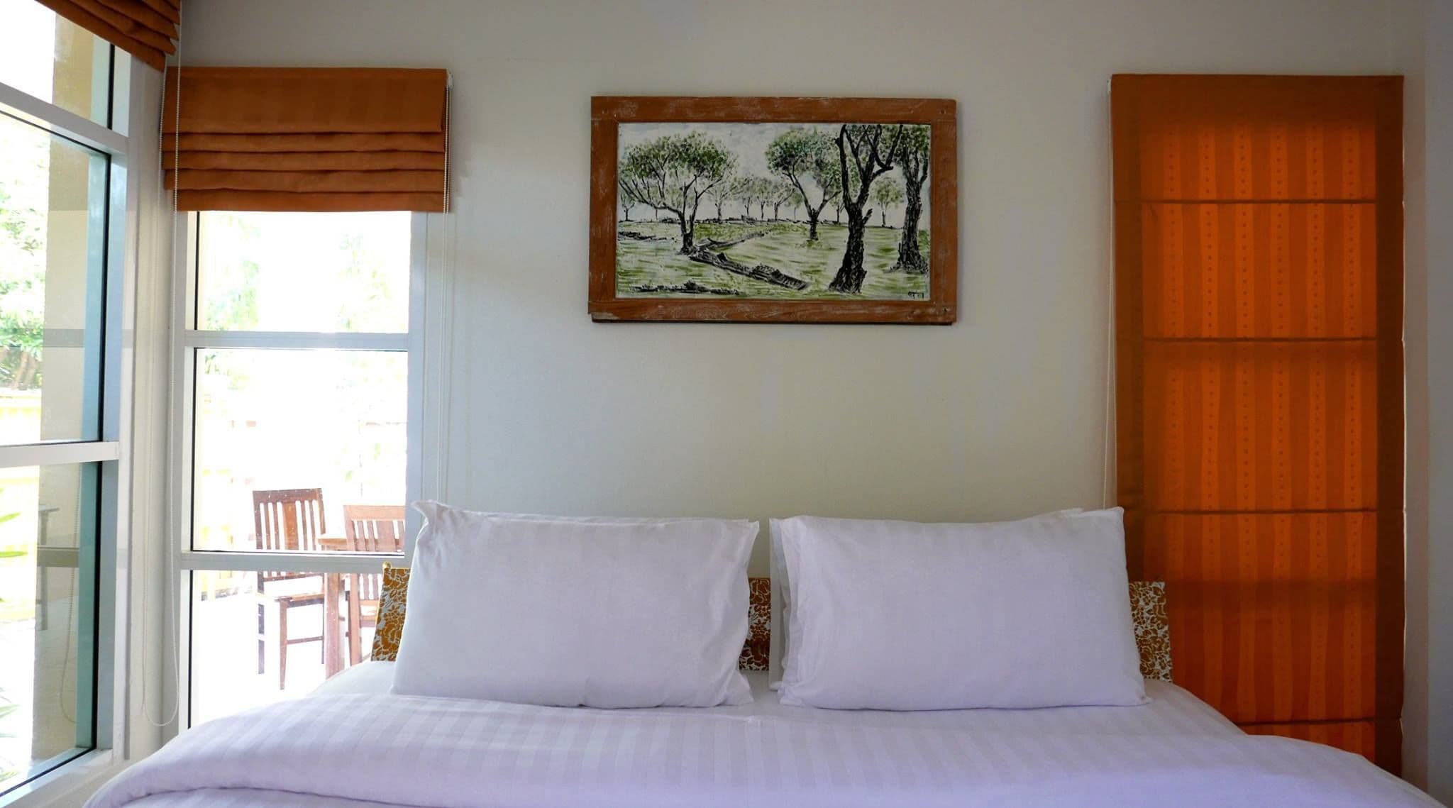 A bedroom with a bed, two pillows, a window with blinds, a painting of trees, and a wooden wardrobe.