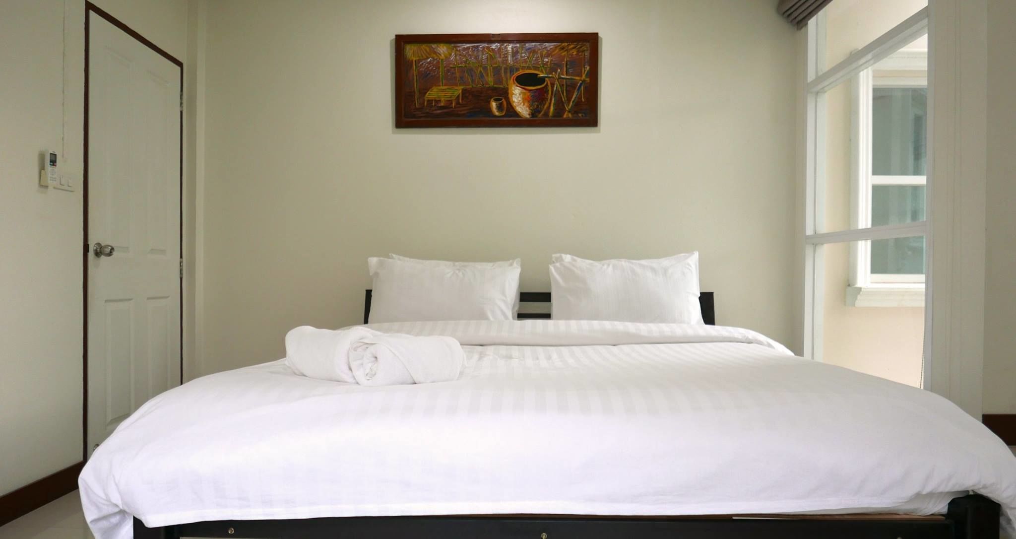 A neatly made bed with white linens, a painting above it, and a window on the right.