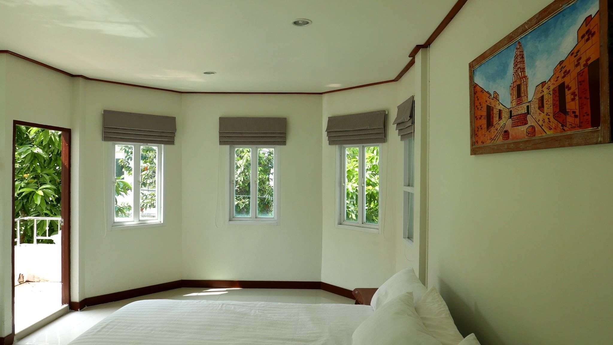 A bedroom with a large bed, three windows with blinds, a ceiling fan, and a painting of a building on the wall.