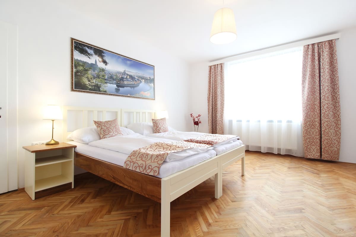 A hotel room with a wooden floor, a bed with white bedding, a nightstand with a lamp, a painting of a cityscape, and patterned curtains.