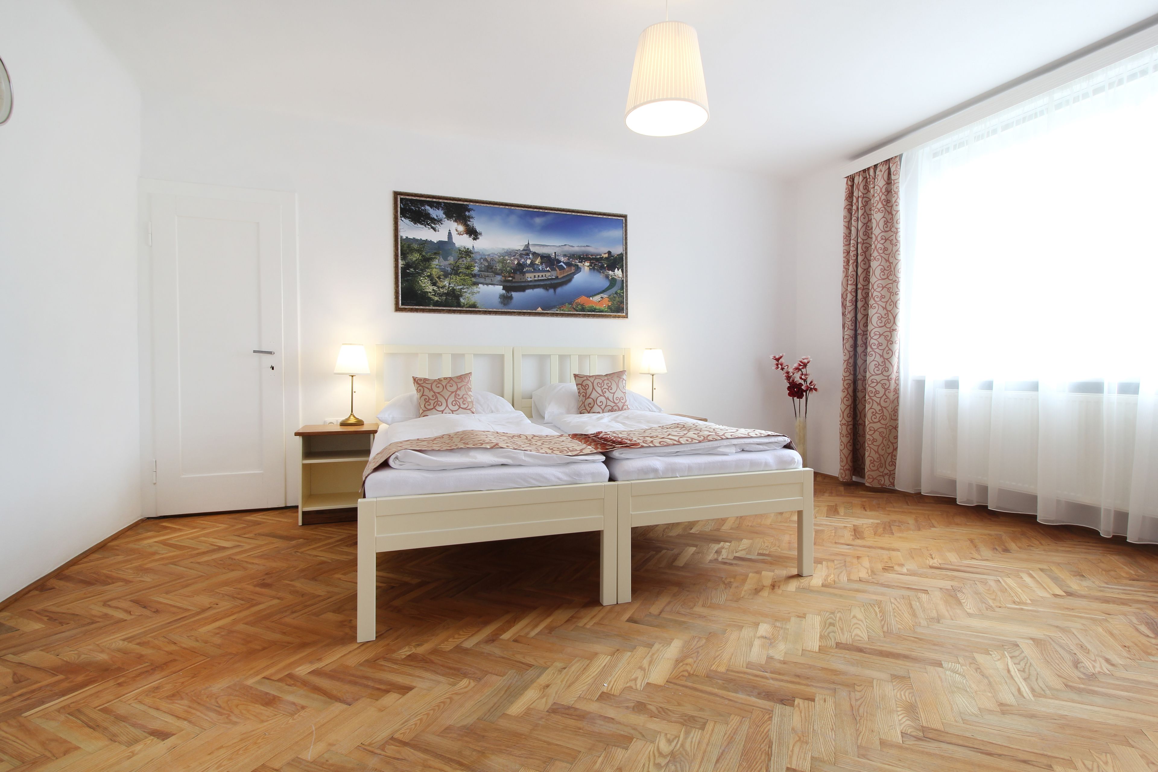 Une chambre avec un parquet en bois disposé en motif à chevrons, un lit blanc avec deux oreillers, une table de chevet avec une lampe et un tableau représentant un paysage accroché au mur.