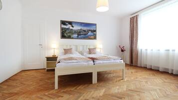 Une chambre avec un parquet en bois disposé en motif à chevrons, un lit blanc avec deux oreillers, une table de chevet avec une lampe et un tableau représentant un paysage accroché au mur.
