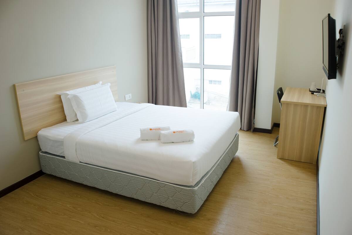 A neatly made bed with white linens and two towels, a wooden headboard, a small wooden desk with a chair, and a window with curtains.