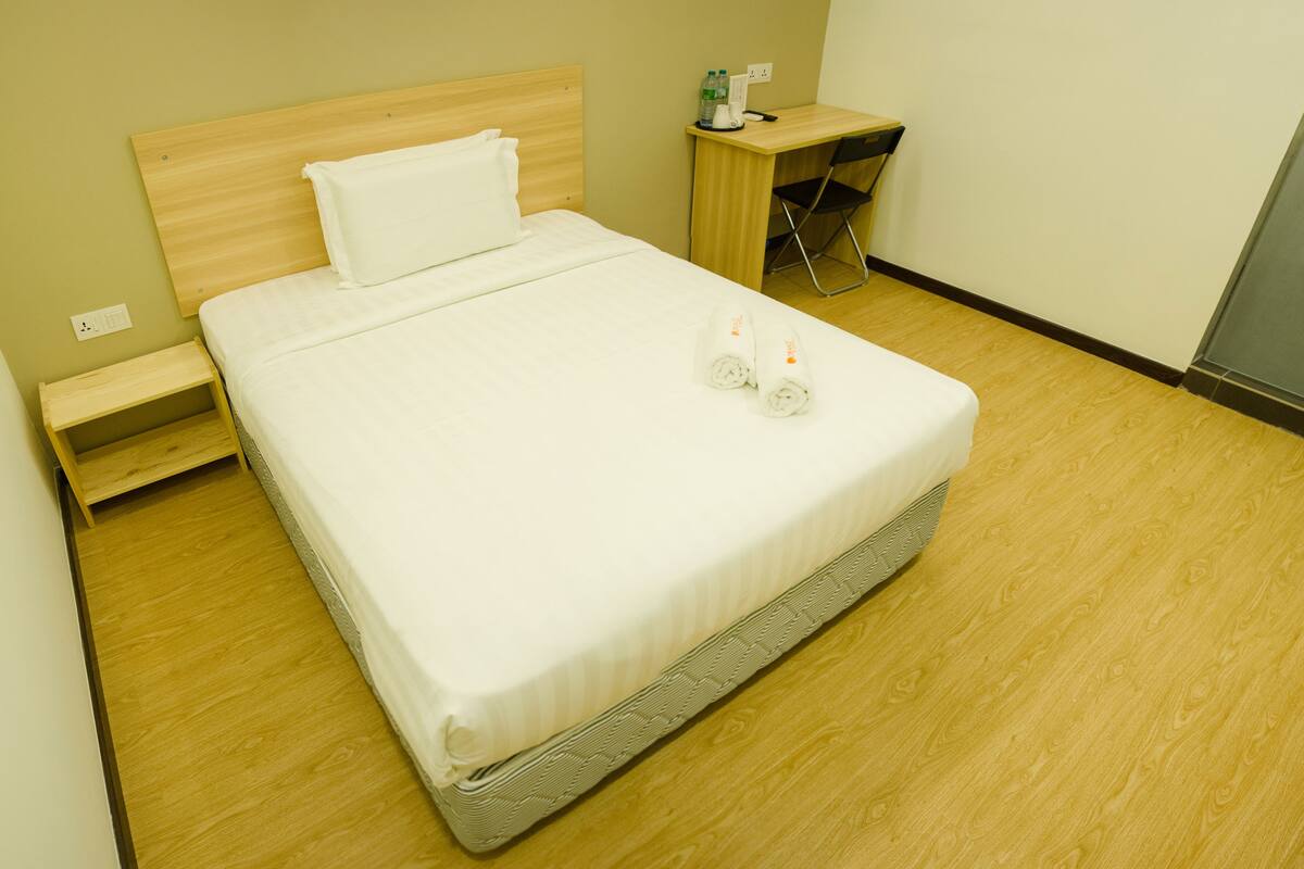 A neatly made bed with white linens, a wooden headboard, a small wooden nightstand with a lamp, and a chair.