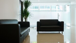 A modern lobby with two black sofas, a large potted plant, and a glass door leading to a bright area with large windows. - Orange Hotels Sri Petaling (Kuala Lumpur)
