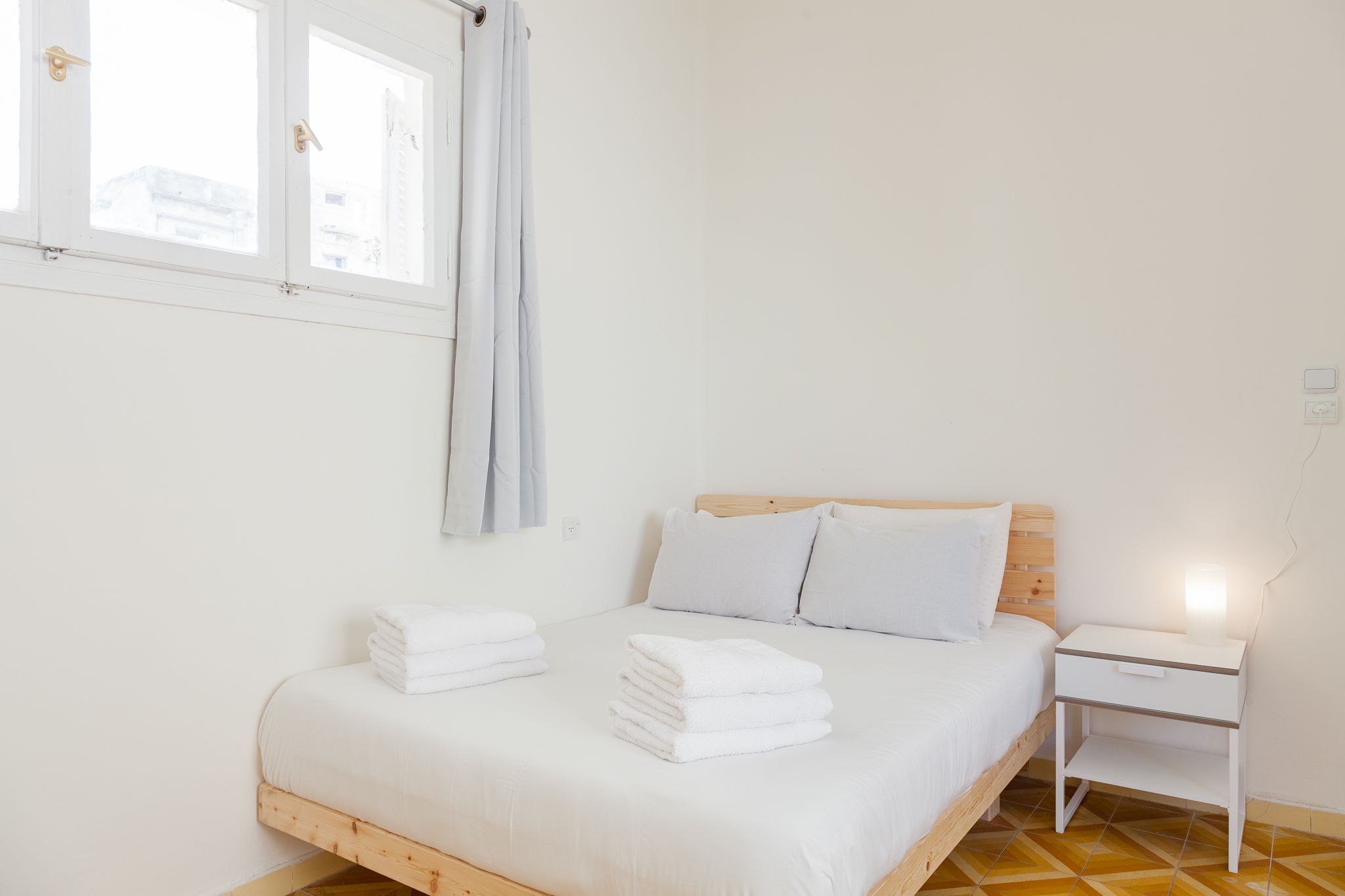 Une chambre à coucher avec un lit en bois, du linge de lit blanc, une table de chevet avec une lampe et une fenêtre avec des rideaux.