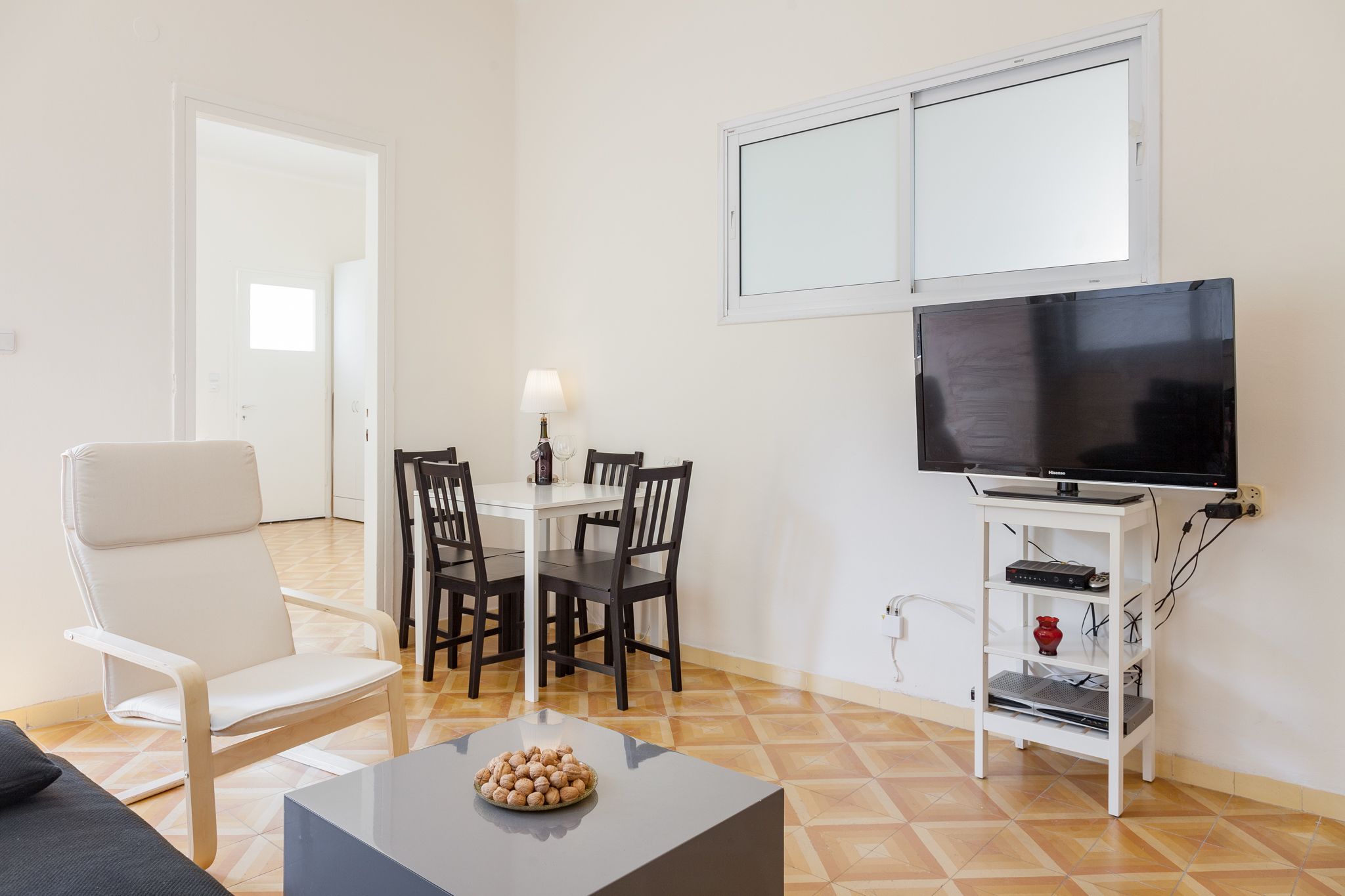 Una sala de estar con un sofá blanco, una mesa de comedor con sillas, un televisor sobre un mueble blanco y un piso de baldosas.