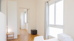 Hotelkamer met een bed, nachtkastje, een klein krukje, een raam met gordijnen en een gang naar een andere kamer.