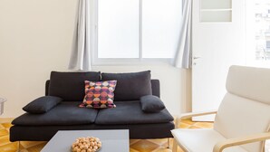 Een moderne woonkamer met een donkergrijze bank, een kleurrijk kussen, een witte fauteuil en een kleine tafel met een schaal lekkernijen.