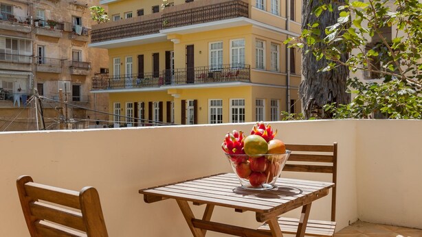 Een balkon met een houten tafel en stoelen, een schaal fruit en uitzicht op meerlaags gebouwen.