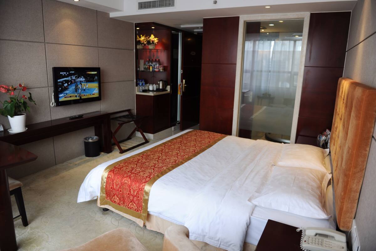 A hotel room with a large bed, a desk with a chair, a television mounted on the wall, and a small potted plant.
