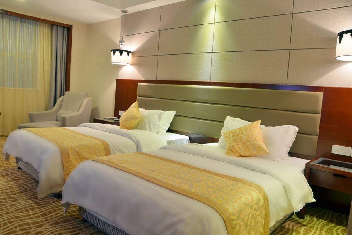 A hotel room with three beds, each with white bedding and yellow accents, a wooden headboard, and a nightstand with a lamp.