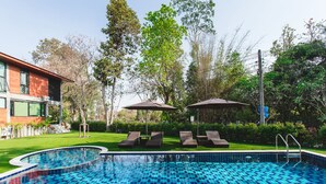 A swimming pool with lounge chairs and umbrellas, surrounded by greenery and a building. - Stucco Loft Residency (Chiang Mai)