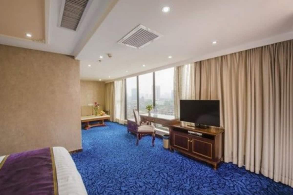 A hotel room with a large bed, a desk with a chair, a television, and a city view through large windows.