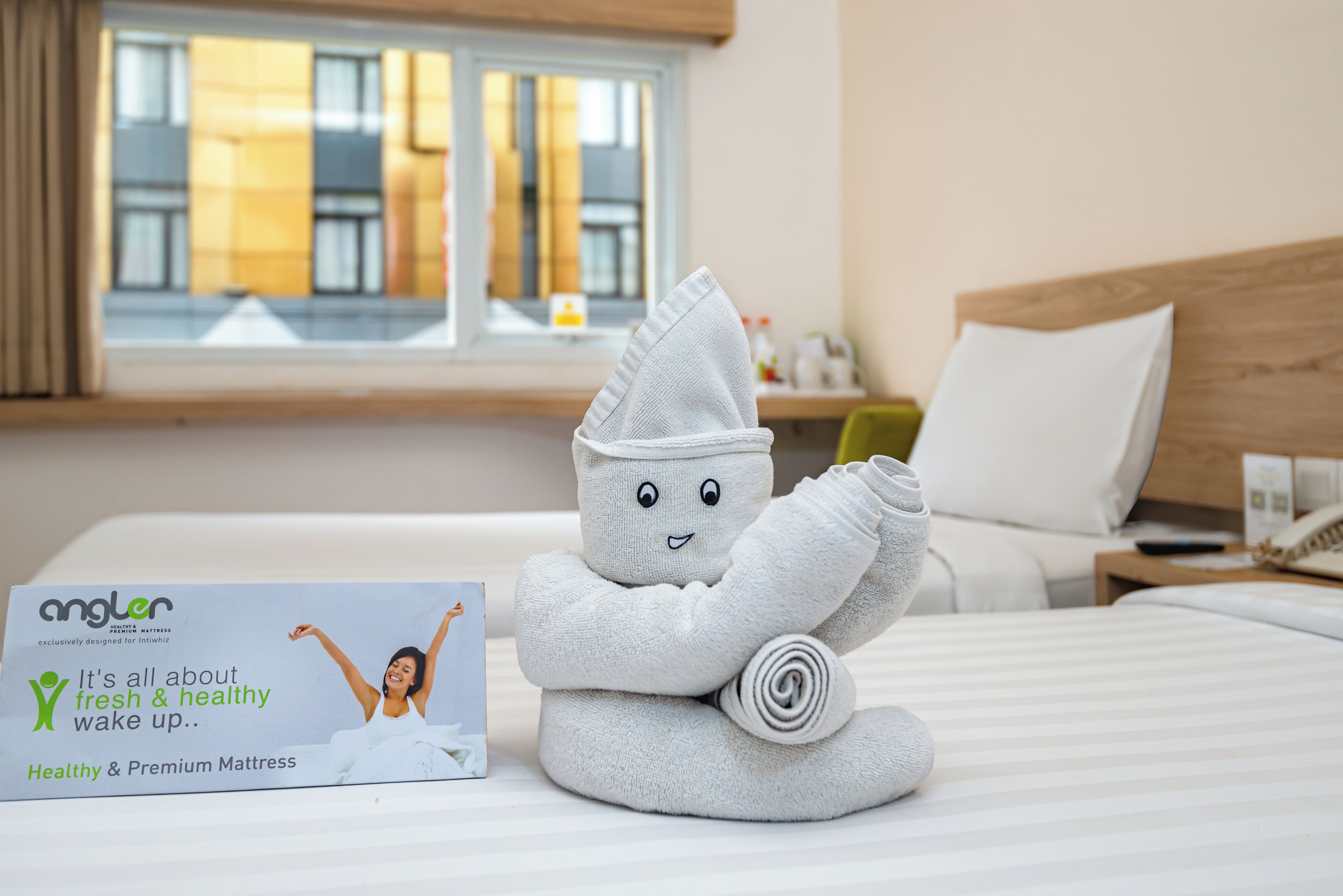 Una habitación de hotel con una cama bien hecha, una ventana con cortinas, una mesita de noche con lámpara y una escultura de toalla.