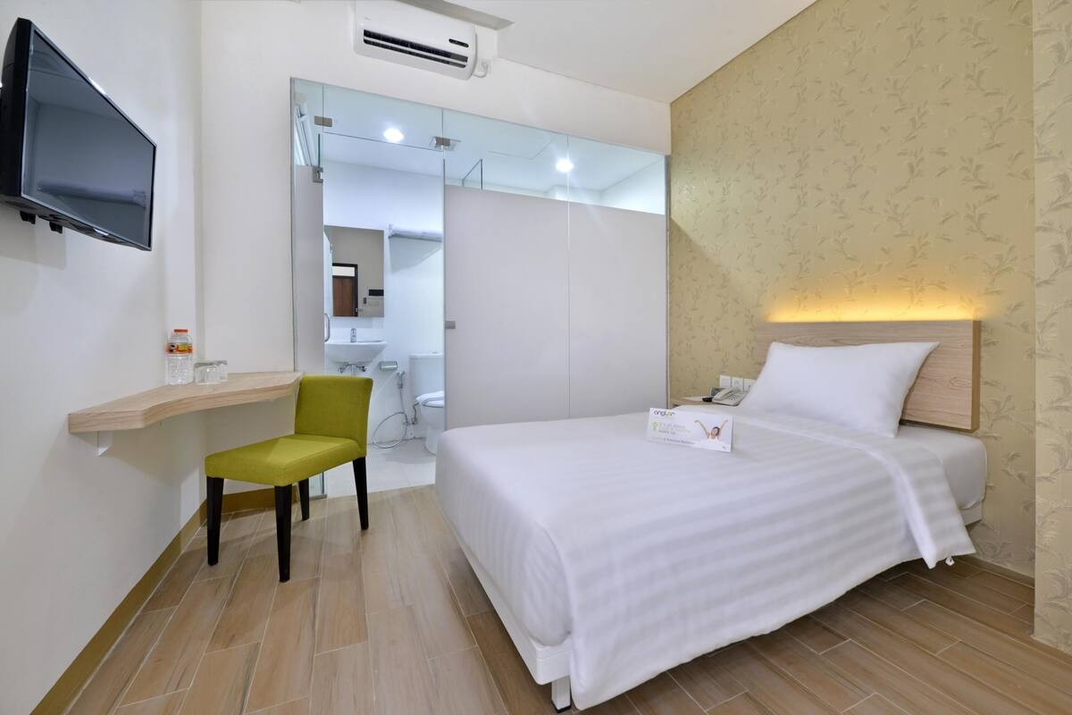 A hotel room with a bed, a desk with a chair, a TV, and a bathroom visible through an open door.