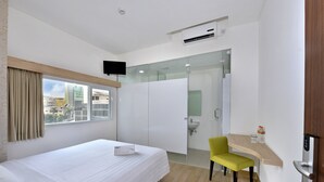 Habitación de hotel con cama, una silla, escritorio con computadora, baño con lavamanos y televisor montado en la pared.
