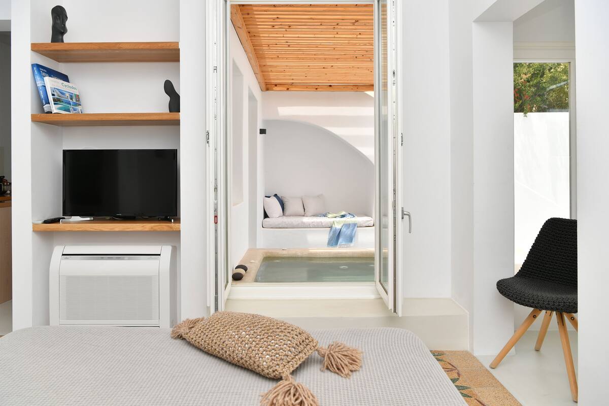 Un dormitorio moderno con televisión, estantería, cama y un rincón de descanso con vistas al exterior.