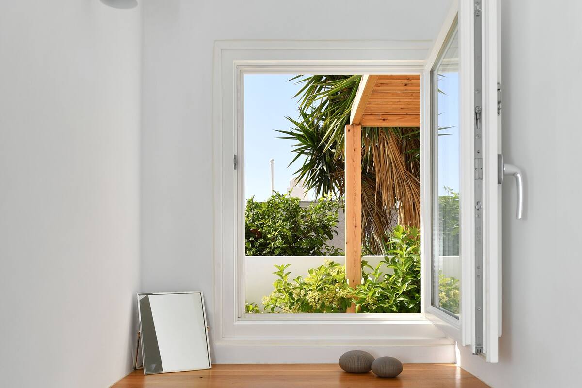 Habitación de esquina con un escritorio de madera, un pequeño banco, una ventana con persiana abierta y vistas a la vegetación.