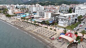 An aerial view of a coastal city with a beach lined with sun loungers and umbrellas, adjacent to multi-story buildings with balconies and a variety of shops and restaurants.