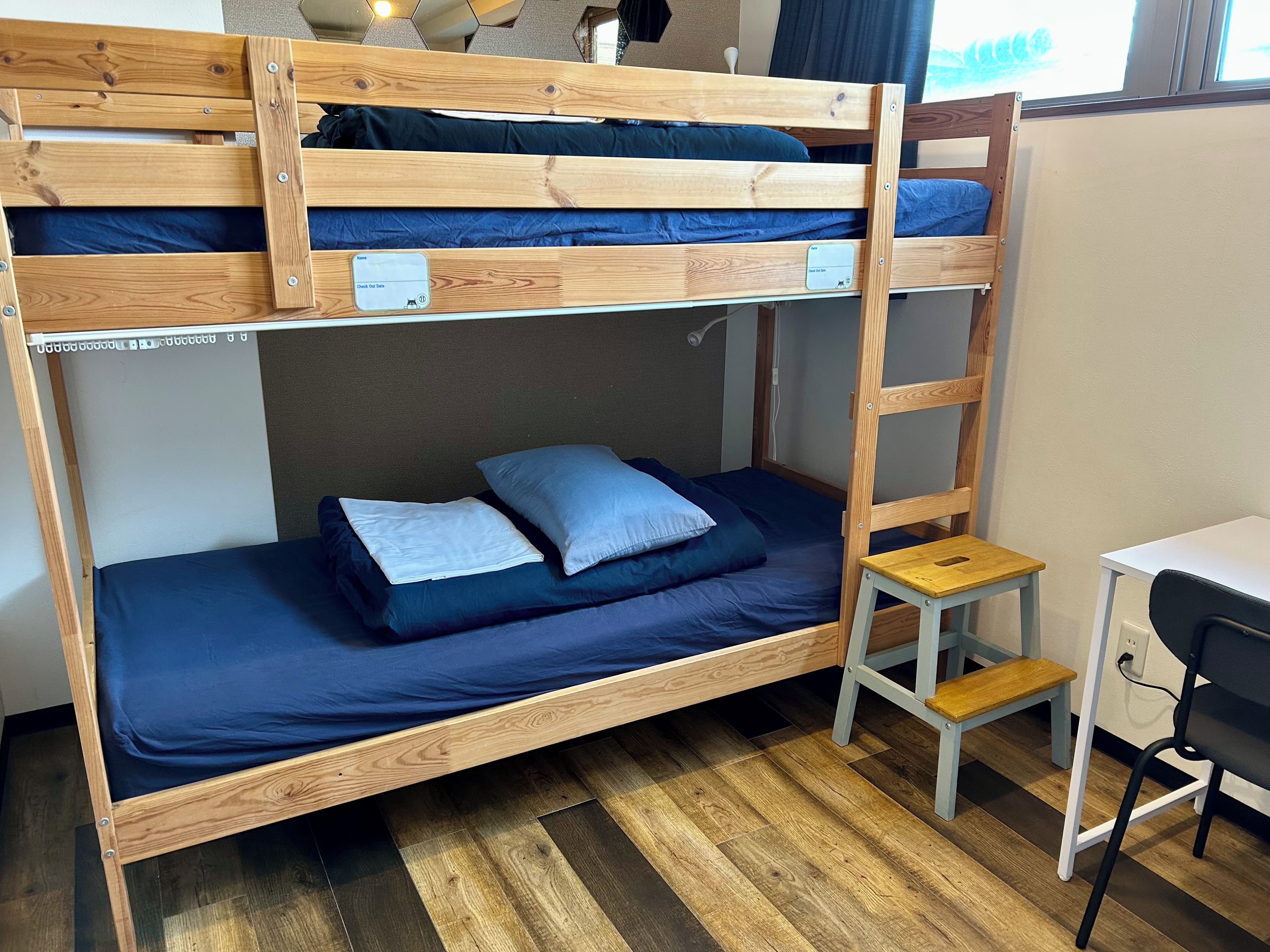 Ein Stockbett mit Schreibtisch und Stuhl, ein kleiner Holzschemel und ein Fenster mit Vorhängen.