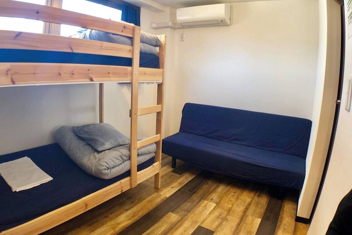 A bunk bed with a ladder, a single bed, and a sofa in a room with wooden flooring and an air conditioning unit on the wall.